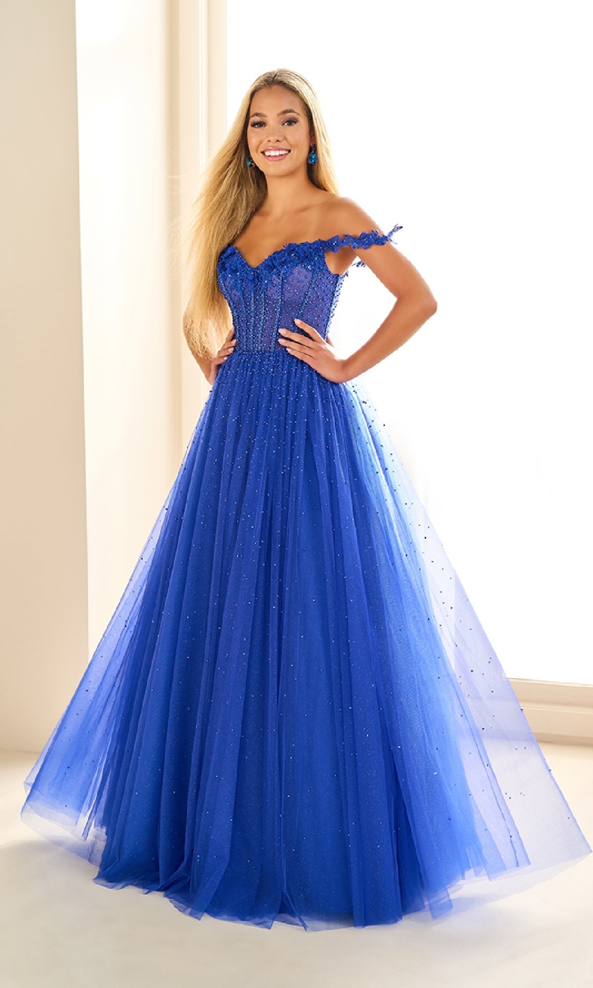 A woman poses indoors in the Ellie Wilde EW36071 long prom dress, an off-the-shoulder gown with a fitted bodice and glitter tulle skirt detailed with small beaded embellishments.